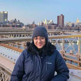 A smiling young woman with brown skin, wearing glasses, a parka and beanie hat, leaning on a bridge within a cityscape..