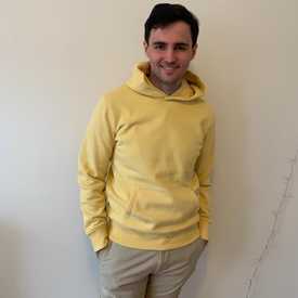 A smiling young man with brown hair appears indoors wearing a yellow hoodie.