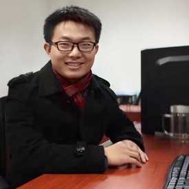 A smiling young Asian man at a desk with short dark hair and glasses.