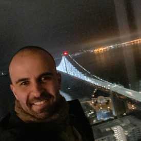 A smiling man appears with a bridge in the background, night time setting.