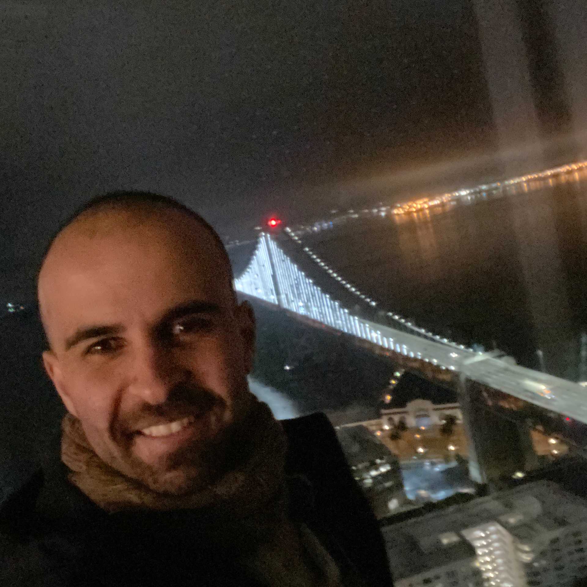 A smiling man appears with a bridge in the background, night time setting.
