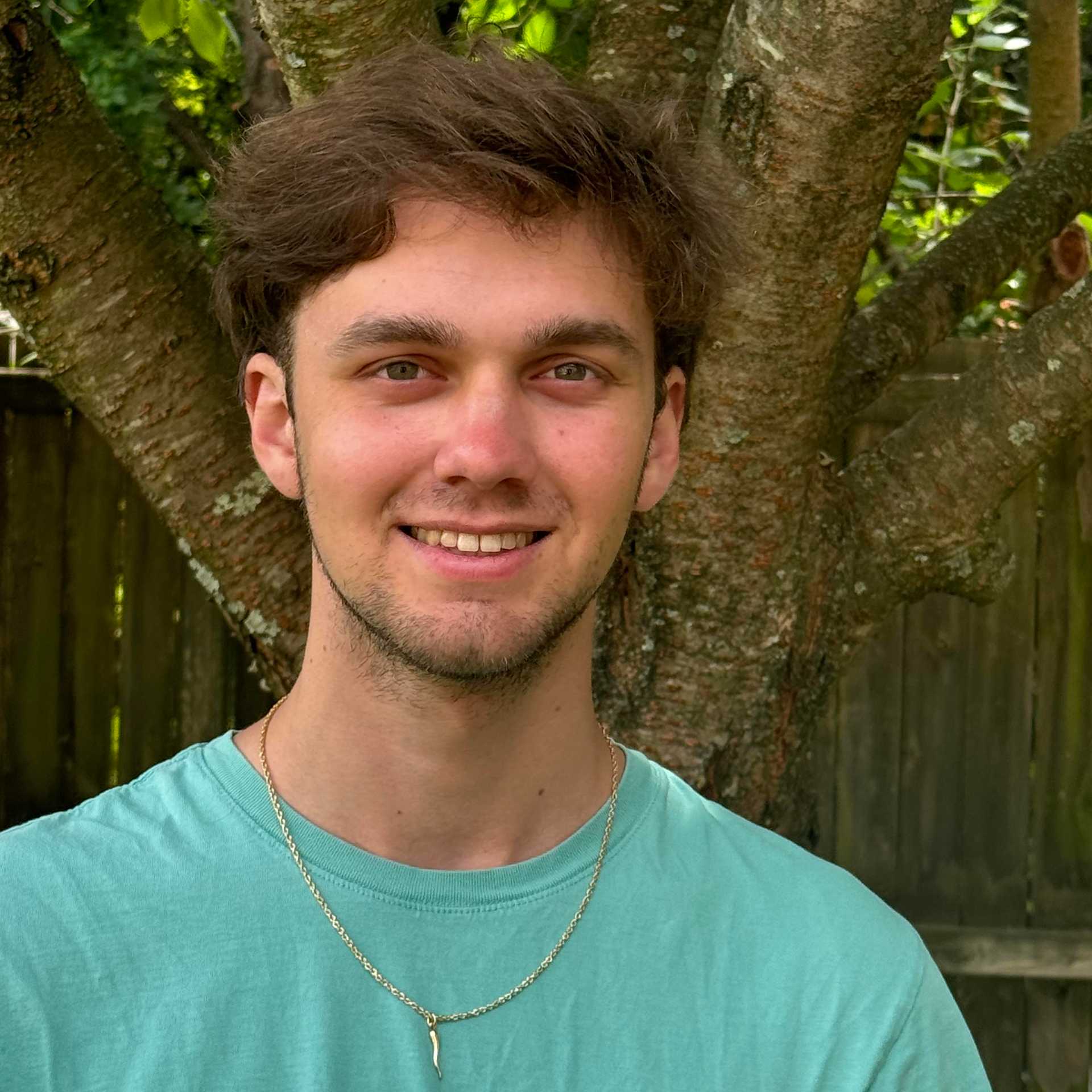 A smiling young man with light wearing a green teeshirt is pictured outdoors, with a tree in the background.