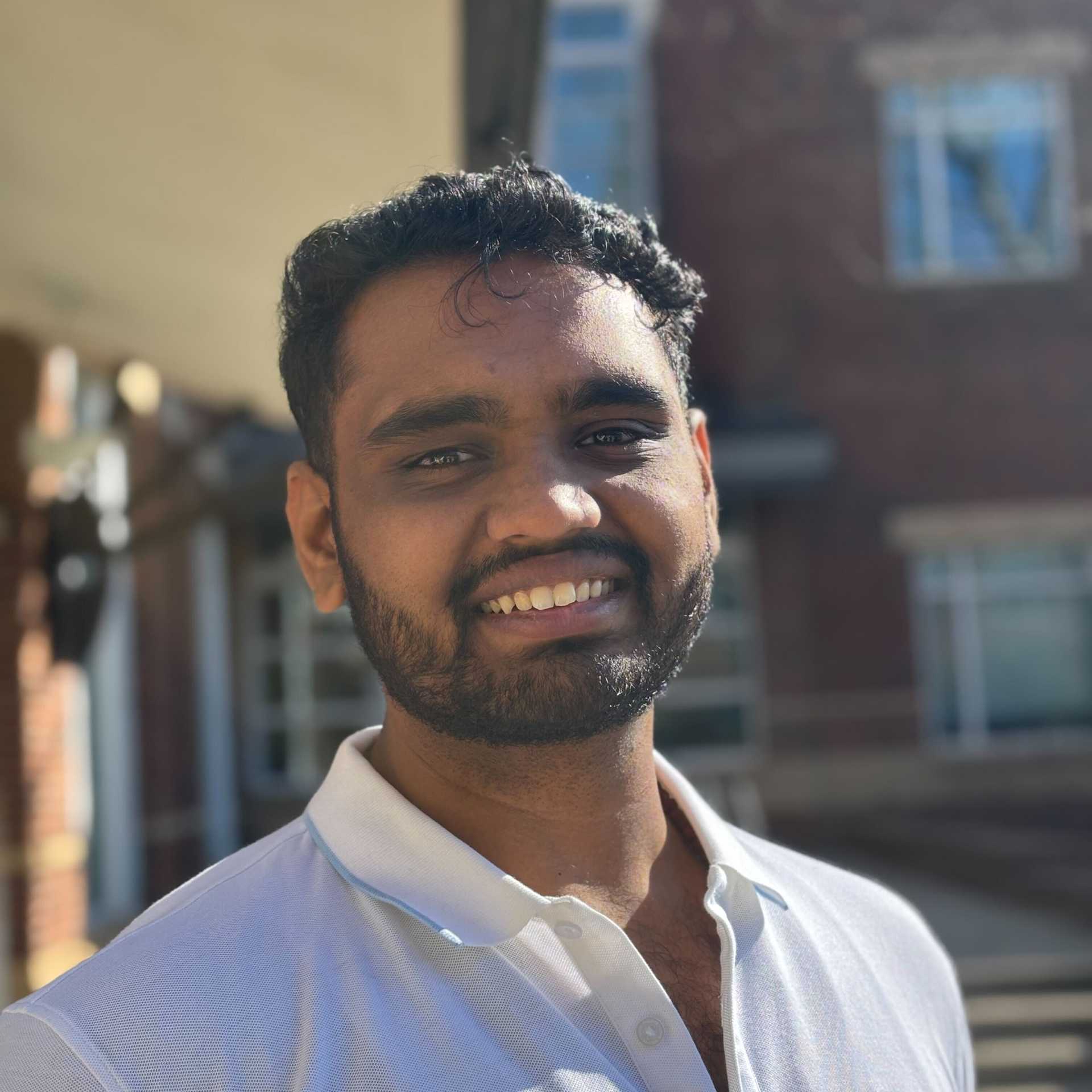 A young man with dark hair and a beard appears outdoors in an urban setting.