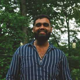 A darl-haired, bearded young man in the outdoors appears smiling; 