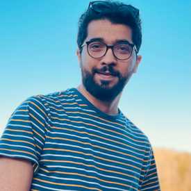 A smiling, bearded young man wearing glasses and a striped shirt appears outdoors with a blue sky background.