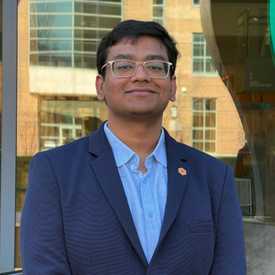 A smiling young man appears wearing glasses and a blue suit coat.