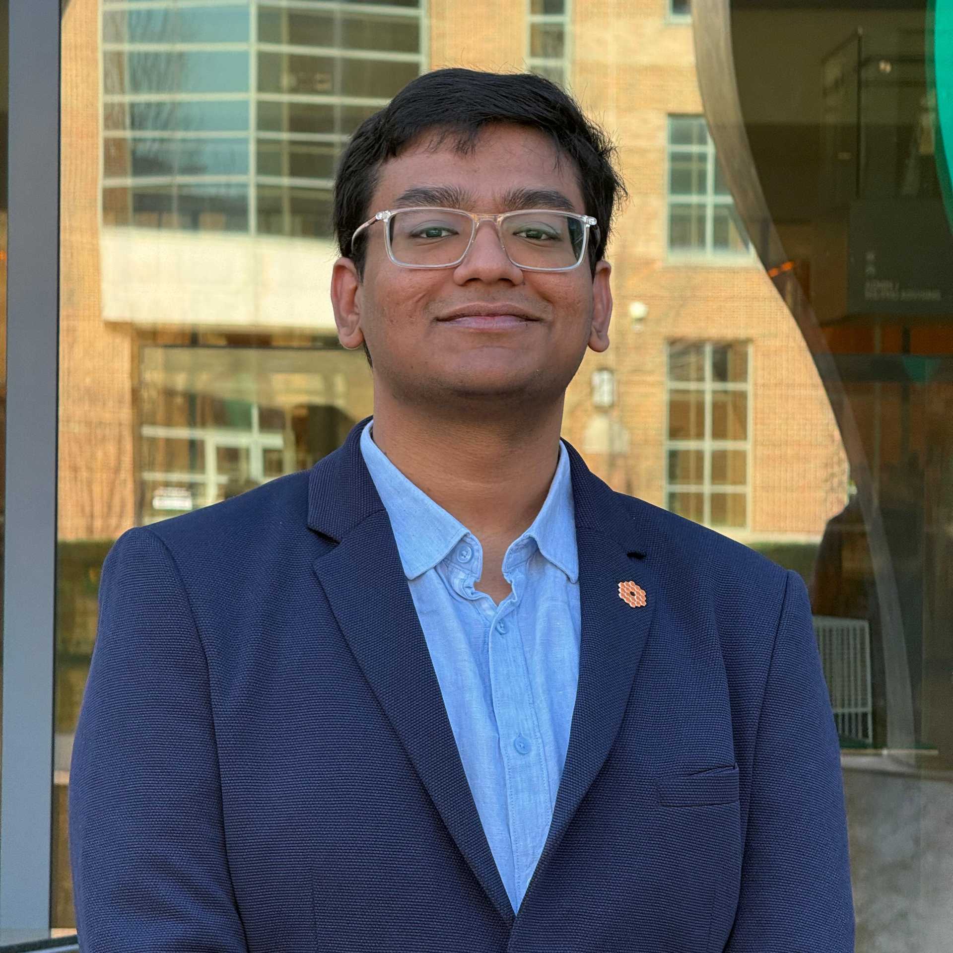 A smiling young man appears wearing glasses and a blue suit coat.