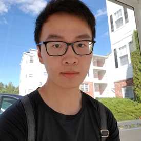 A photo of Kaixin Huang, who looks intently at the camera. He wears a black t-shirt and backpack. The sky is blue and cloudy behind him.