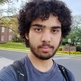 A young man appears outdoors, wearing a backpack and blue teeshirt. He has dark curly hair, a moustache and beard.