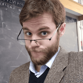 A bearded young man with light brown hair peers over his glasses, with a questioning look, and a blackboard in the background.