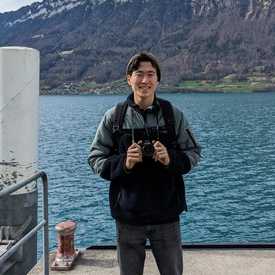 A young man appears outdoors with water and mountains behind him, he is holding a camera.